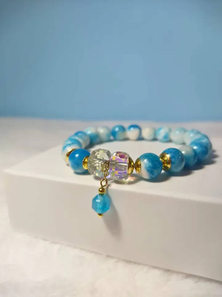 Pulseira de contas azuis e brancas com cristais furta-cor e pingente, sobre uma caixa branca
