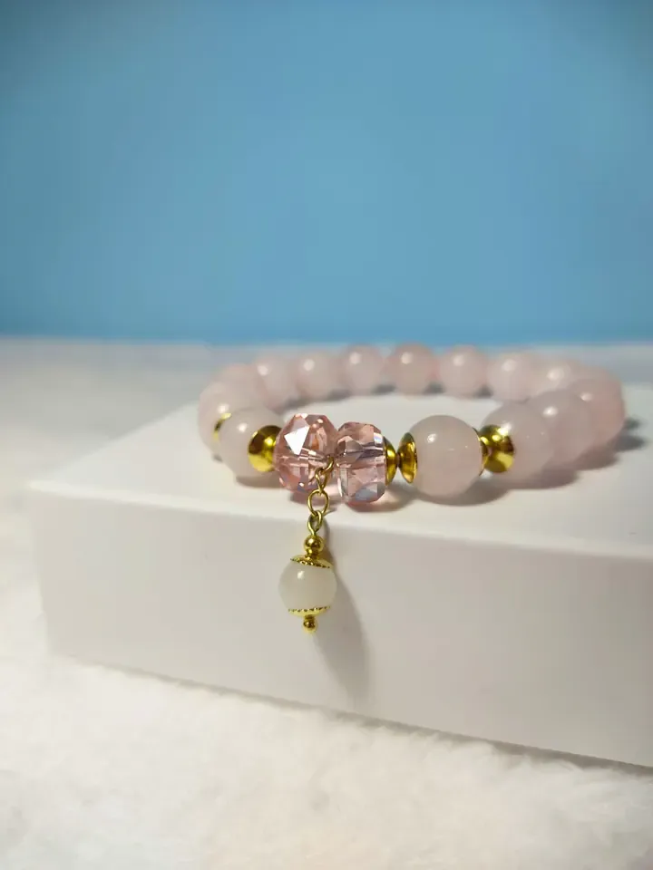 Pulseira de contas rosa-claro com cristais rosa, entremeios dourados e pingente, sobre uma caixa branca